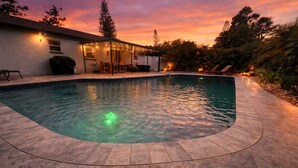 Ein Pool mit geschwungener Kante, umgeben von einer Terrassenfläche mit überdachter Sitzgelegenheit und einem Gebäude im Hintergrund.