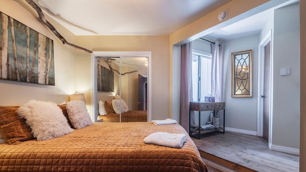 A bedroom with a large bed, a mirror, a wooden floor, and a door leading to another room. - Iconic Lakeview Estate | 4BR | Sleeps 10 (Lake Arrowhead)