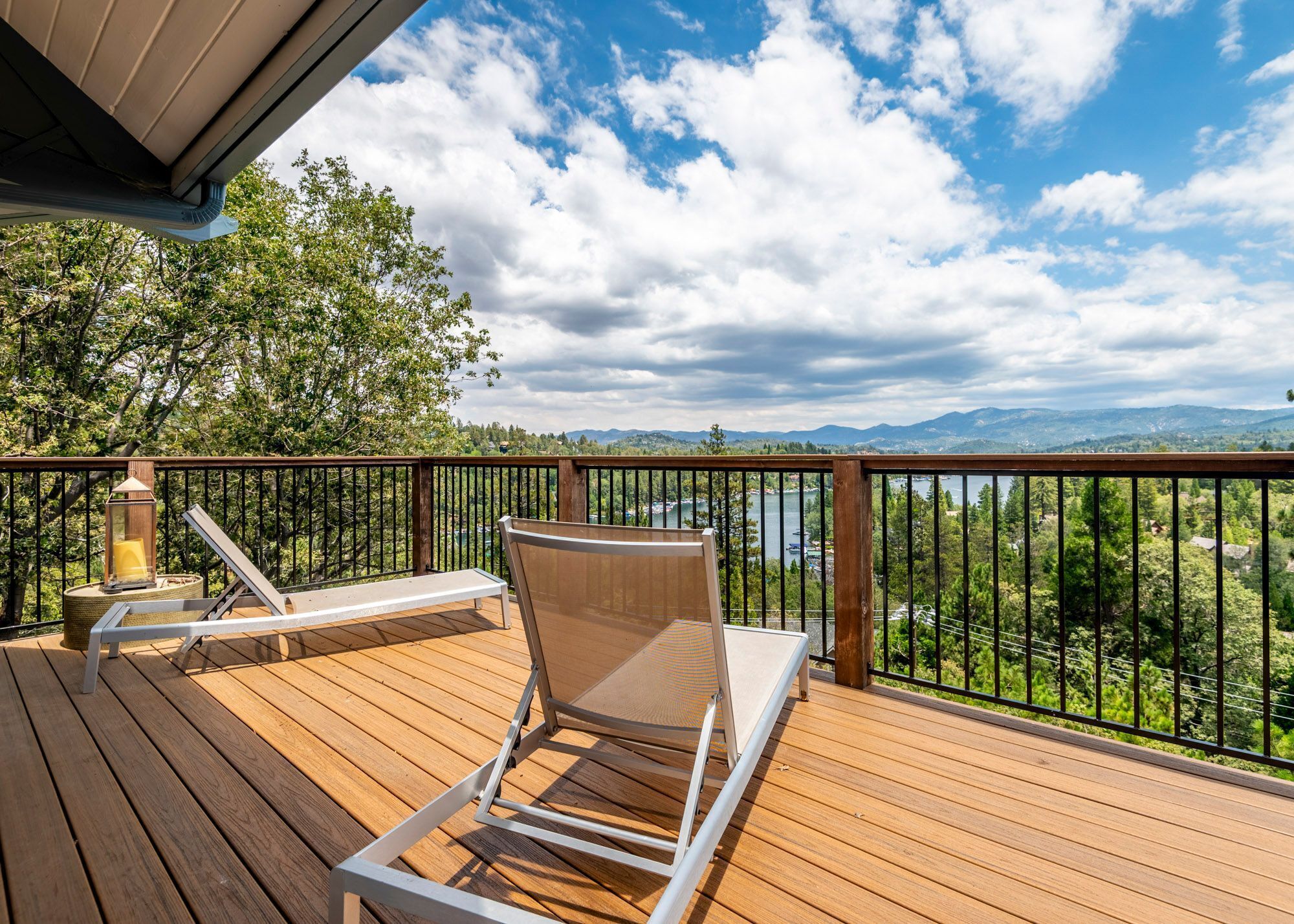 Eine Holzterrasse mit zwei Liegestühlen, einer Laterne und Blick auf einen See und Berge.