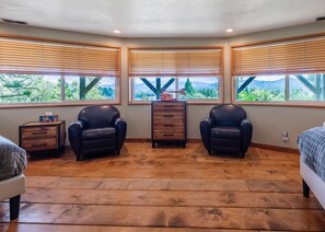 A bedroom with three black leather chairs, a wooden dresser, and a bed. Large windows with wooden blinds offer a view of greenery and hills. - Iconic Lakeview Estate | 4BR | Sleeps 10 (Lake Arrowhead)