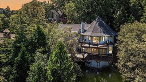 A multi-story house with a balcony, surrounded by dense trees and a body of water. - Iconic Lakeview Estate | 4BR | Sleeps 10 (Lake Arrowhead)