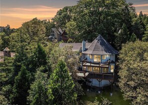 A multi-story house with a balcony, surrounded by dense trees and a body of water. - Iconic Lakeview Estate | 4BR | Sleeps 10 (Lake Arrowhead)