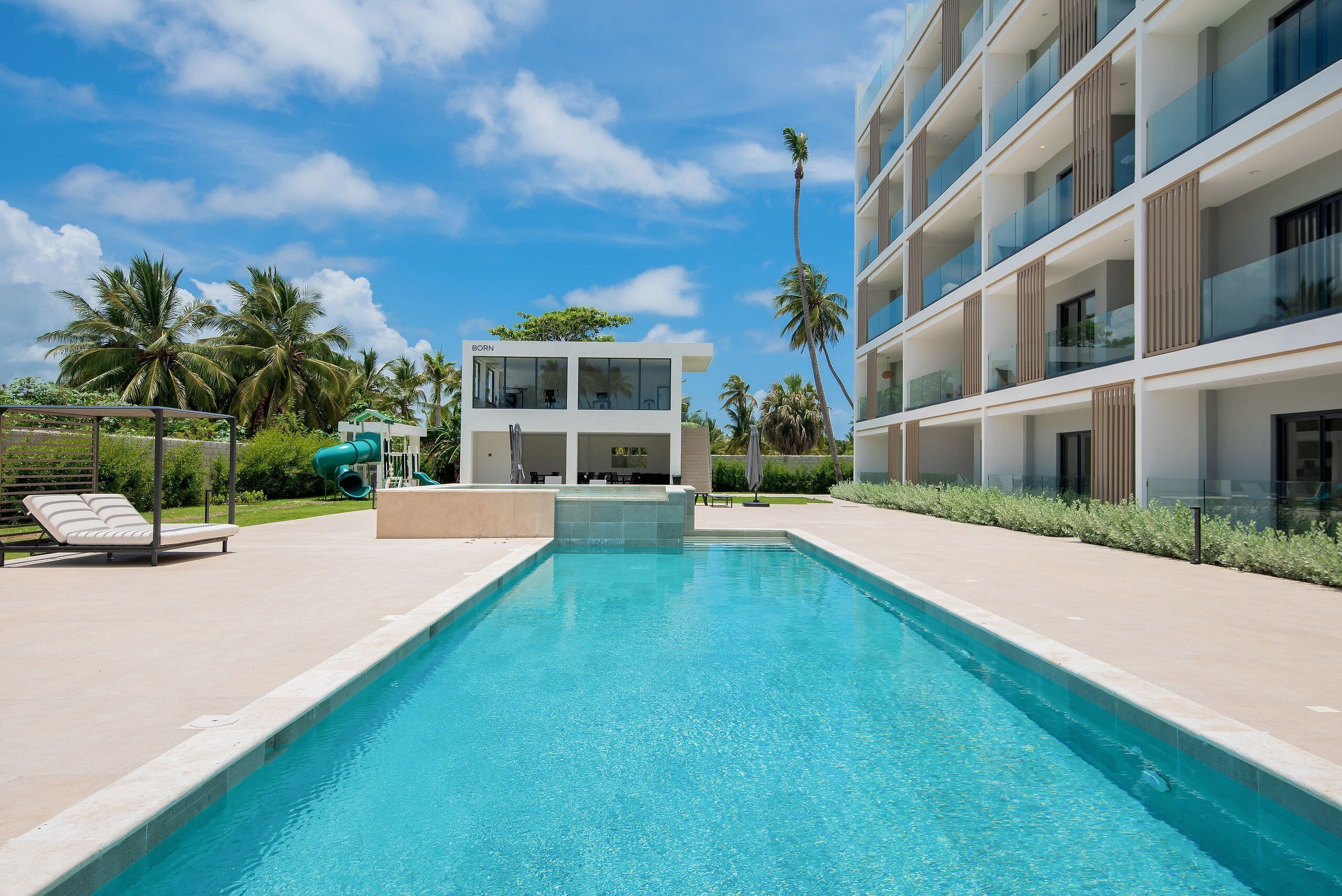 A modern building with a pool, lounge chairs, and palm trees.