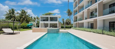 A modern building with a pool, lounge chairs, and palm trees.