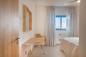 A hotel room with a wooden door, a white desk with a remote control, a single chair, a bed with white bedding, and a window with curtains. - Born | Modern 2BR with Pools and Gym (Bavaro)