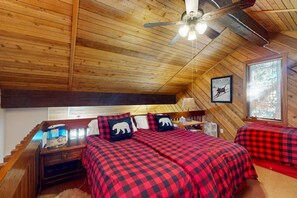 Une chambre de style cabane avec un plafond en pente, un ventilateur de plafond, deux lits avec une literie à carreaux rouges et noirs, une table de chevet, une lampe et un tableau encadré représentant un chien accroché au mur.