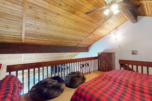 A room with a sloped ceiling, wooden beams, a bed with a red checkered bedspread, a wooden dresser, and a view through a balcony railing. - Cozy Mammoth Condo | Hot Tub & Mountain Adventure (Mammoth Lakes)
