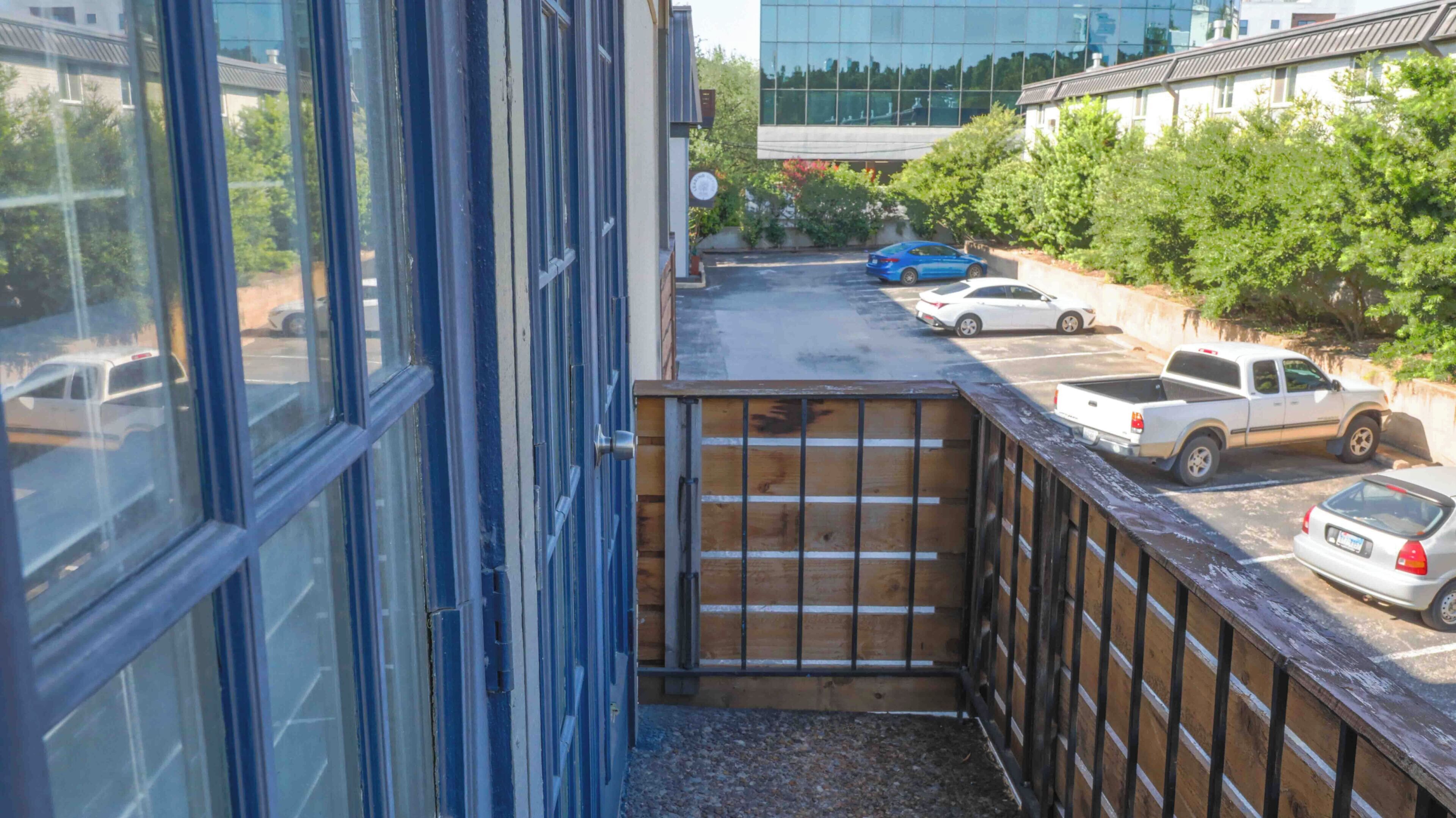Un estrecho pasillo exterior con cerca de madera, una puerta azul y vista a coches estacionados y un edificio al fondo.
