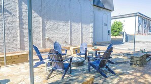 Un espace extérieur aménagé avec des chaises Adirondack et une petite table, un foyer en pierre et un bâtiment en briques blanches portant le numéro 1302.