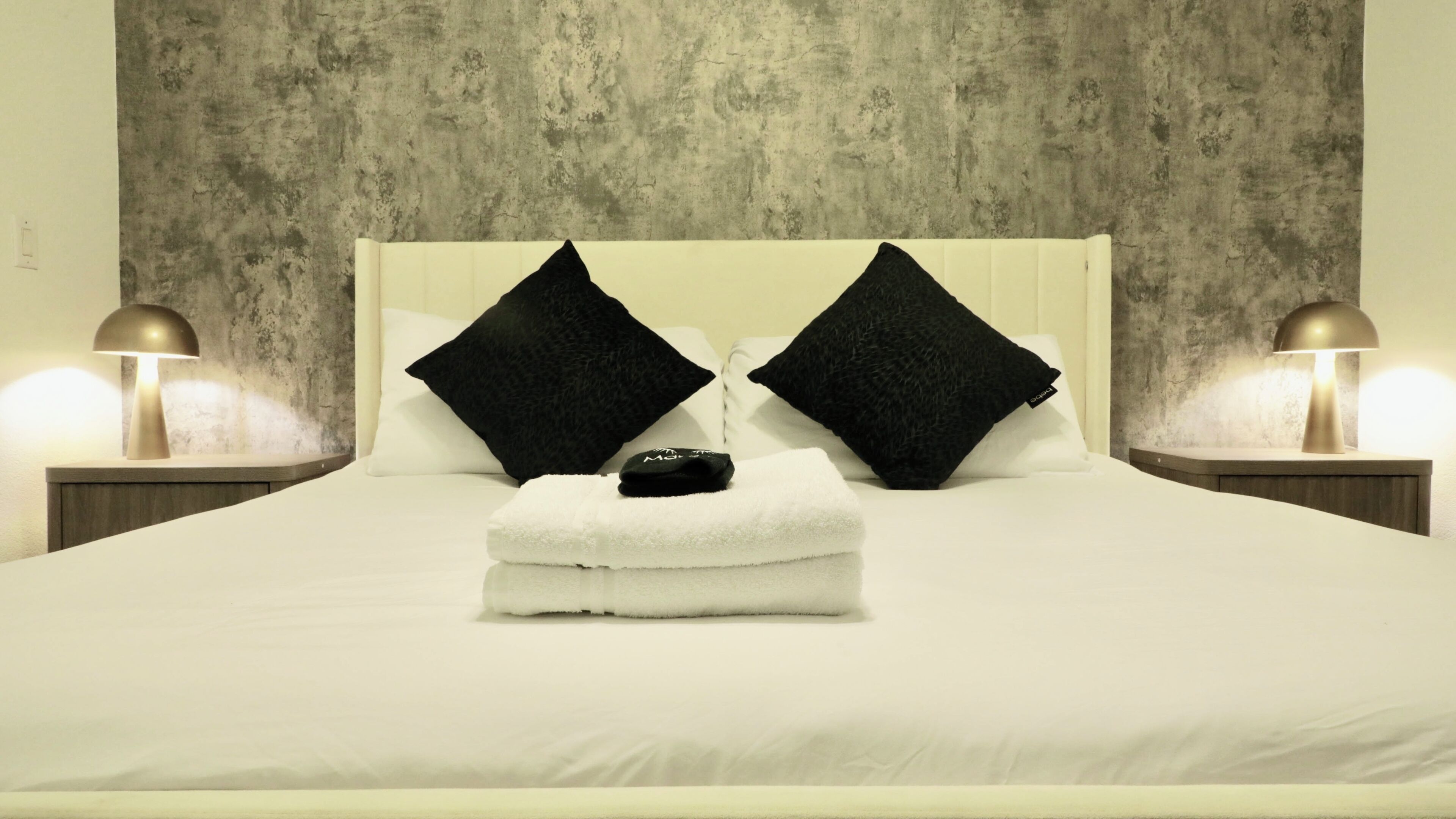 A hotel room with a bed, two black pillows, a white towel, and two bedside lamps.
