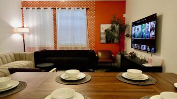 Een woonkamer met een bank, een eettafel gedekt met kommen en borden, een televisie waarop een videogesprek wordt getoond, en een lamp.