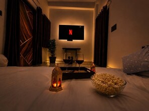 A hotel room with a bed, a TV mounted on the wall, a tray with glasses and a bottle, a bowl of popcorn, and a lit candle. - Luxury 4BHK Private Villa| Pool, Jacuzzi & Home Theater (Liyo Ka Gurha)