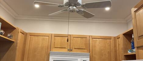 Een keuken met houten kastjes, een witte magnetron, een fornuis en een plafondventilator.