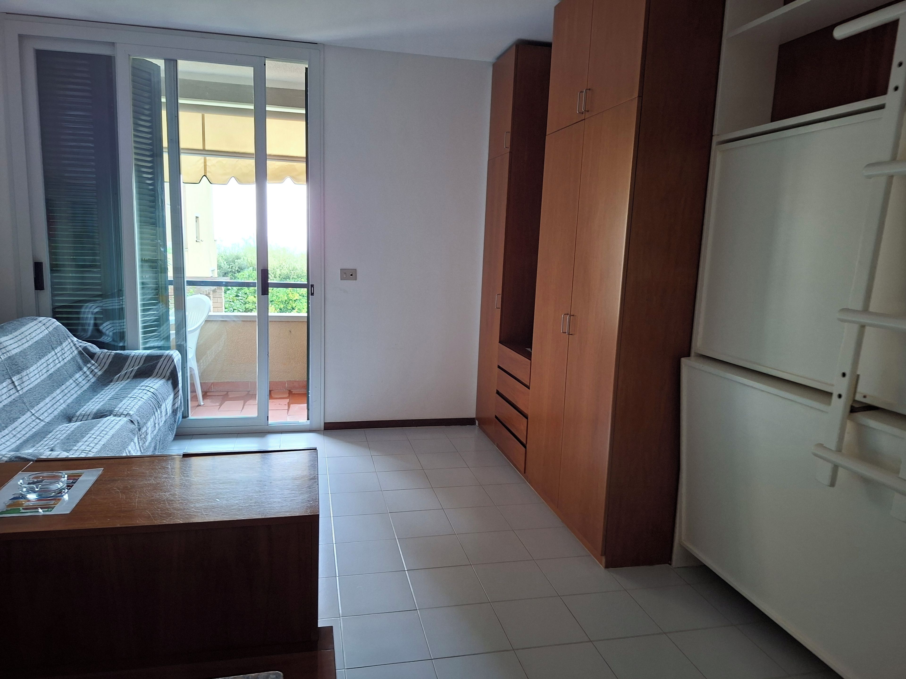 Una stanza con un divano, una porta scorrevole in vetro che dà su un balcone, un armadio in legno e un mobiletto bianco.
