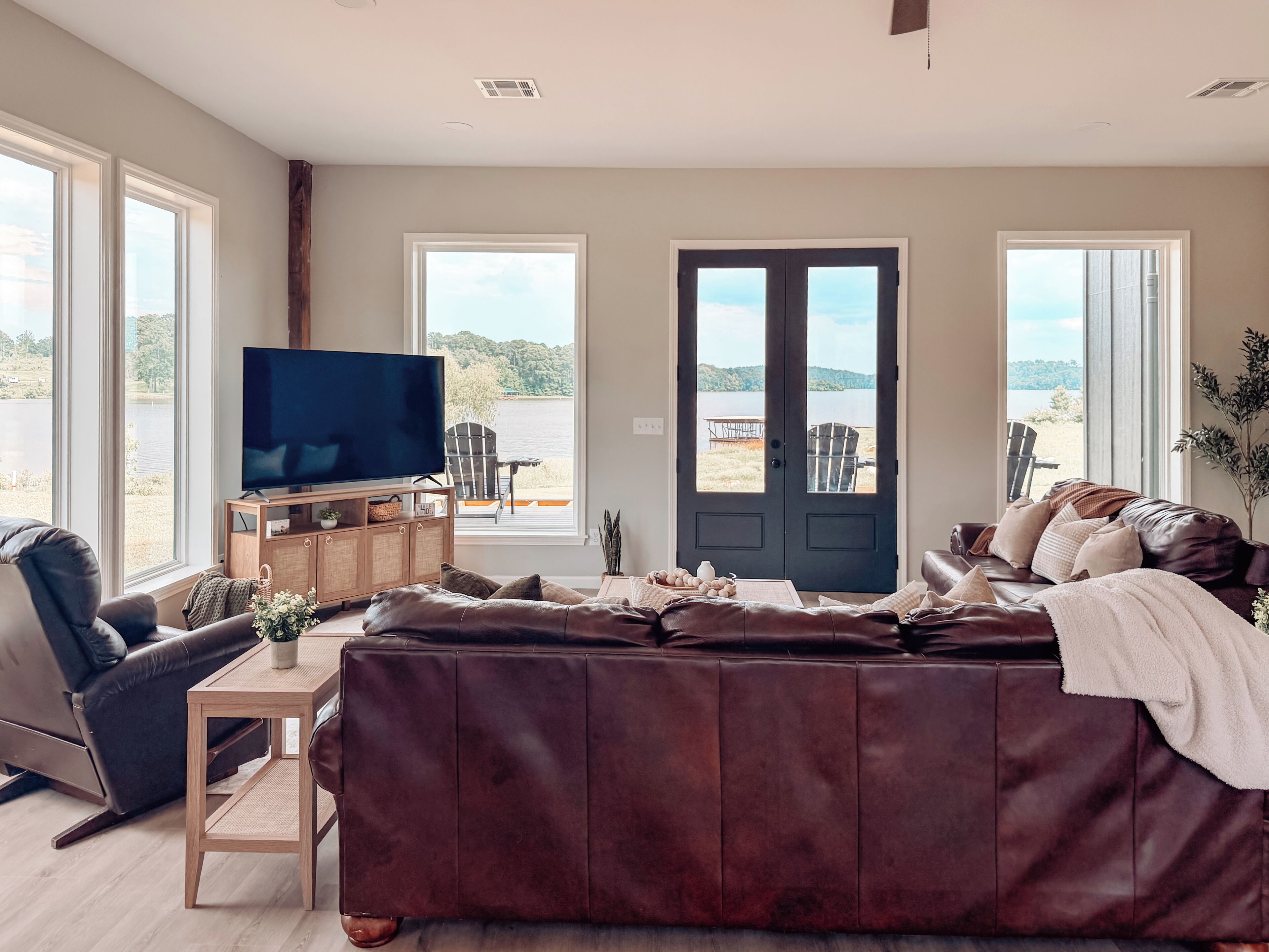 A living room with a large leather sofa, a television, and a view of a lake through a door.
