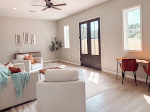 Un salon spacieux comprenant un canapé, un pouf et un bureau avec des chaises. Il y a une grande fenêtre et une porte donnant sur l’extérieur.