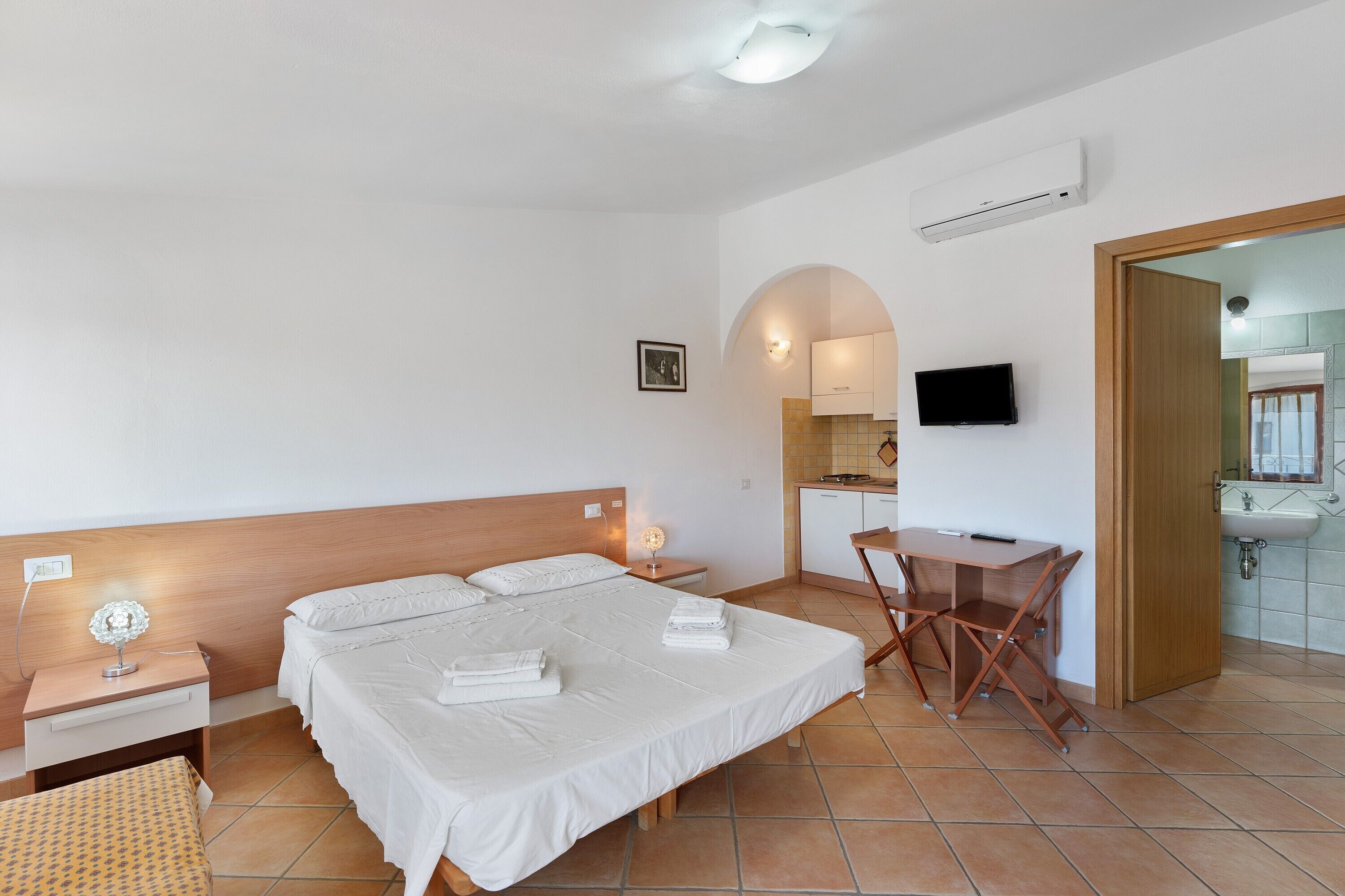 Une chambre d’hôtel comprenant un lit, une table de chevet, une lampe, un bureau, une chaise, une télévision, et donnant sur la cuisine et la salle de bain.