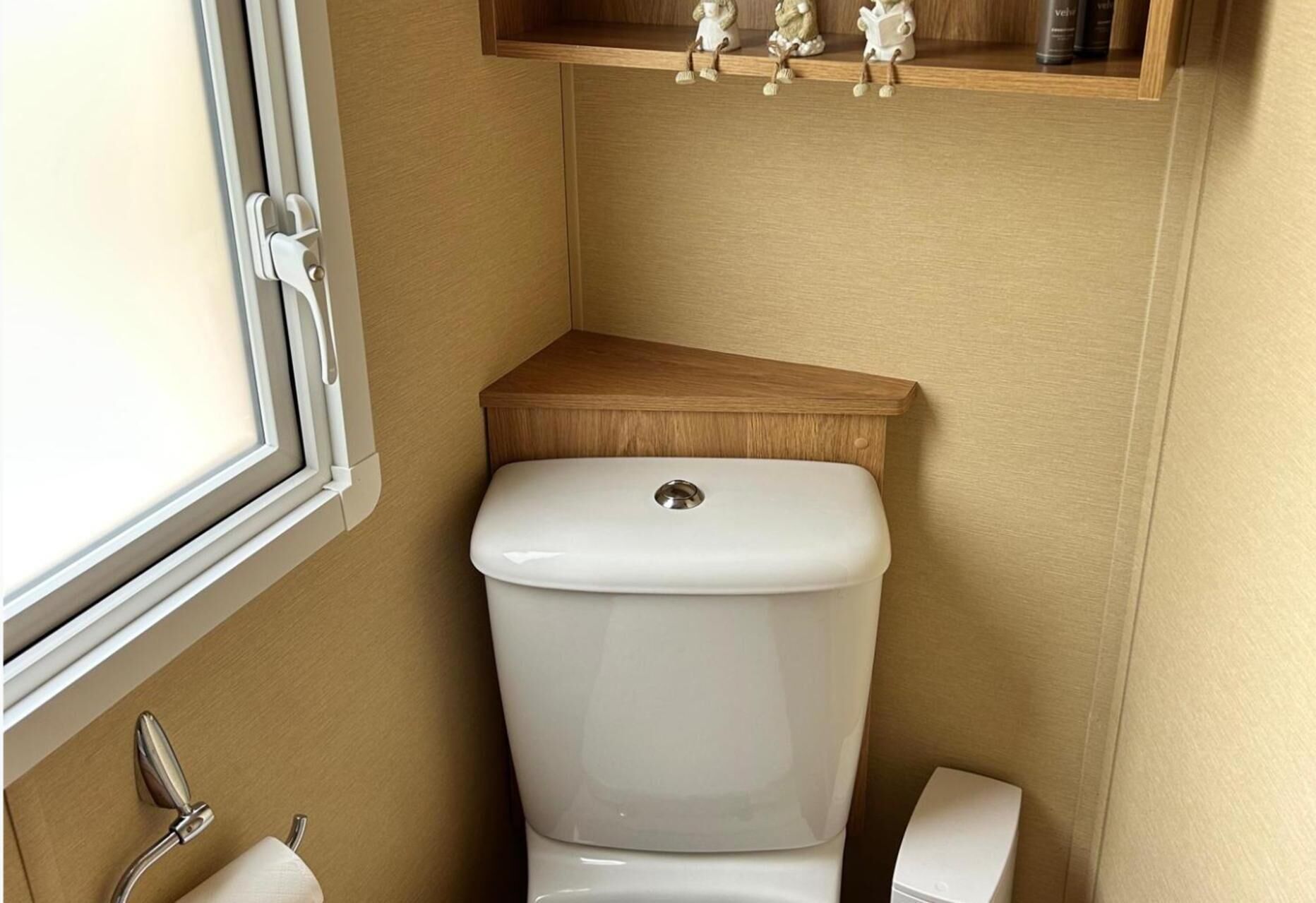 Une salle de bain avec des toilettes blanches, une étagère en bois au-dessus et une fenêtre sur le côté gauche.