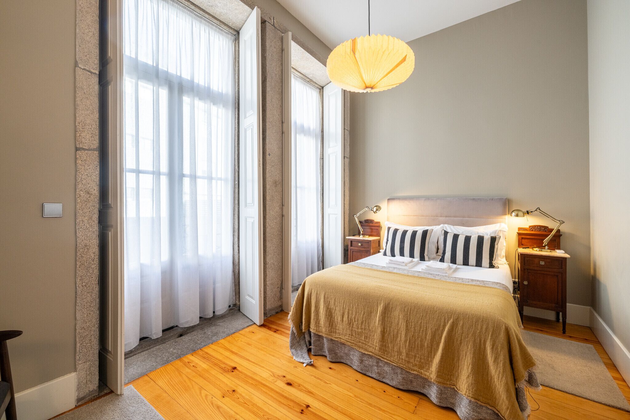 Une chambre à coucher avec un grand lit, une table de chevet, une lampe et une fenêtre ornée de rideaux légers.