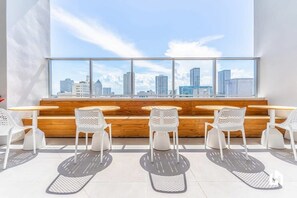 Un intérieur moderne avec vue sur la ville, comprenant un espace salon avec des chaises et des tables blanches, ainsi qu’un banc en bois.