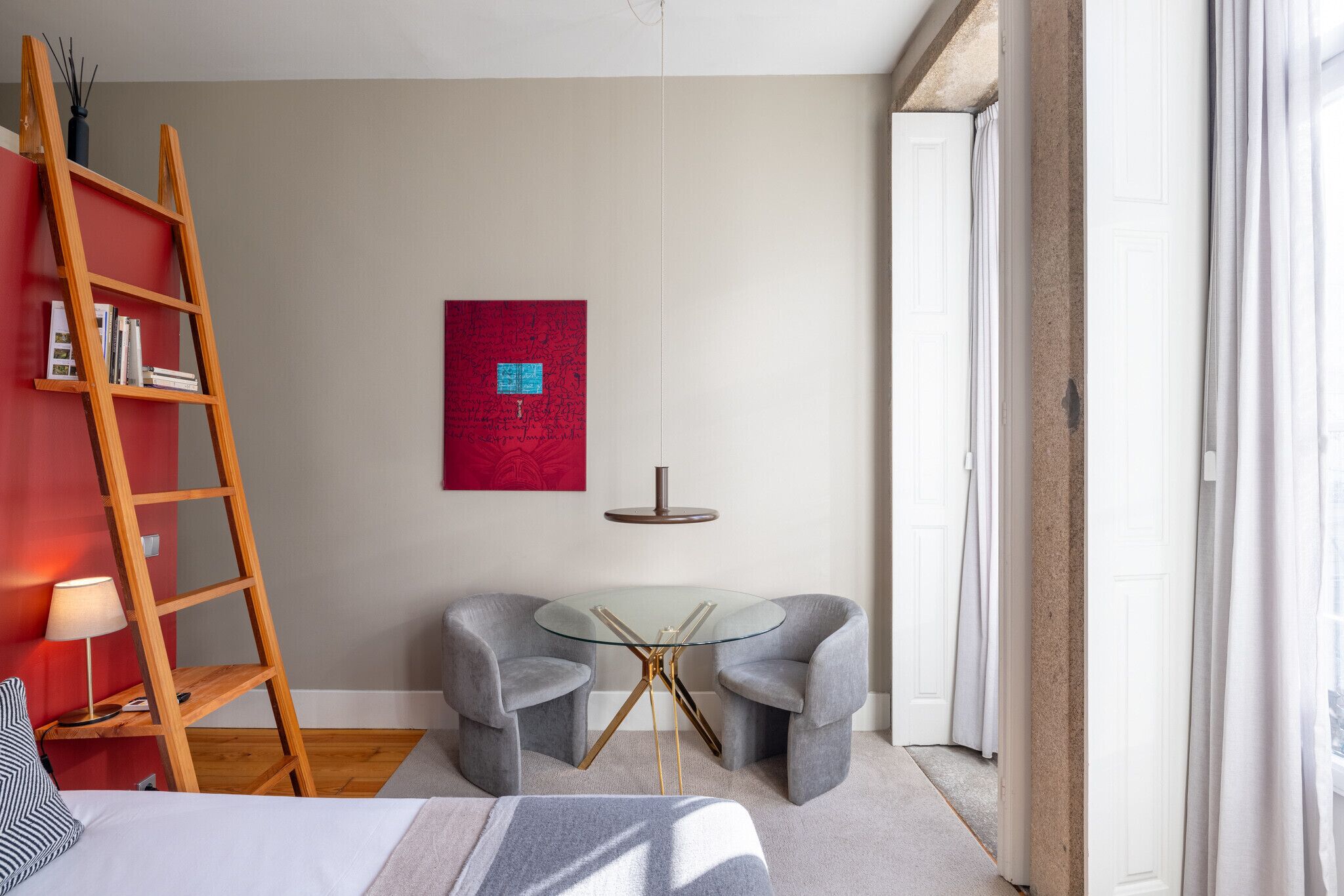 Une chambre moderne avec un lit, un mur rouge, une table en verre avec des chaises, une échelle et un luminaire suspendu.