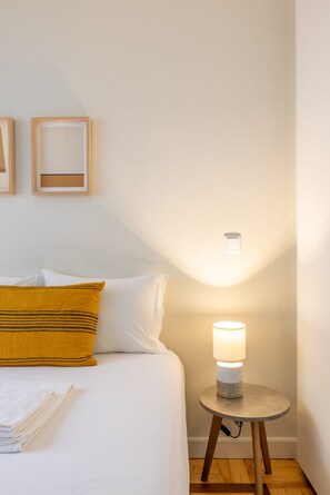 A neatly made bed with white and yellow pillows, a small bedside table with a lamp, and framed pictures on the wall. - Casa do Ofício - Hosty's Gallery Owner (Porto)