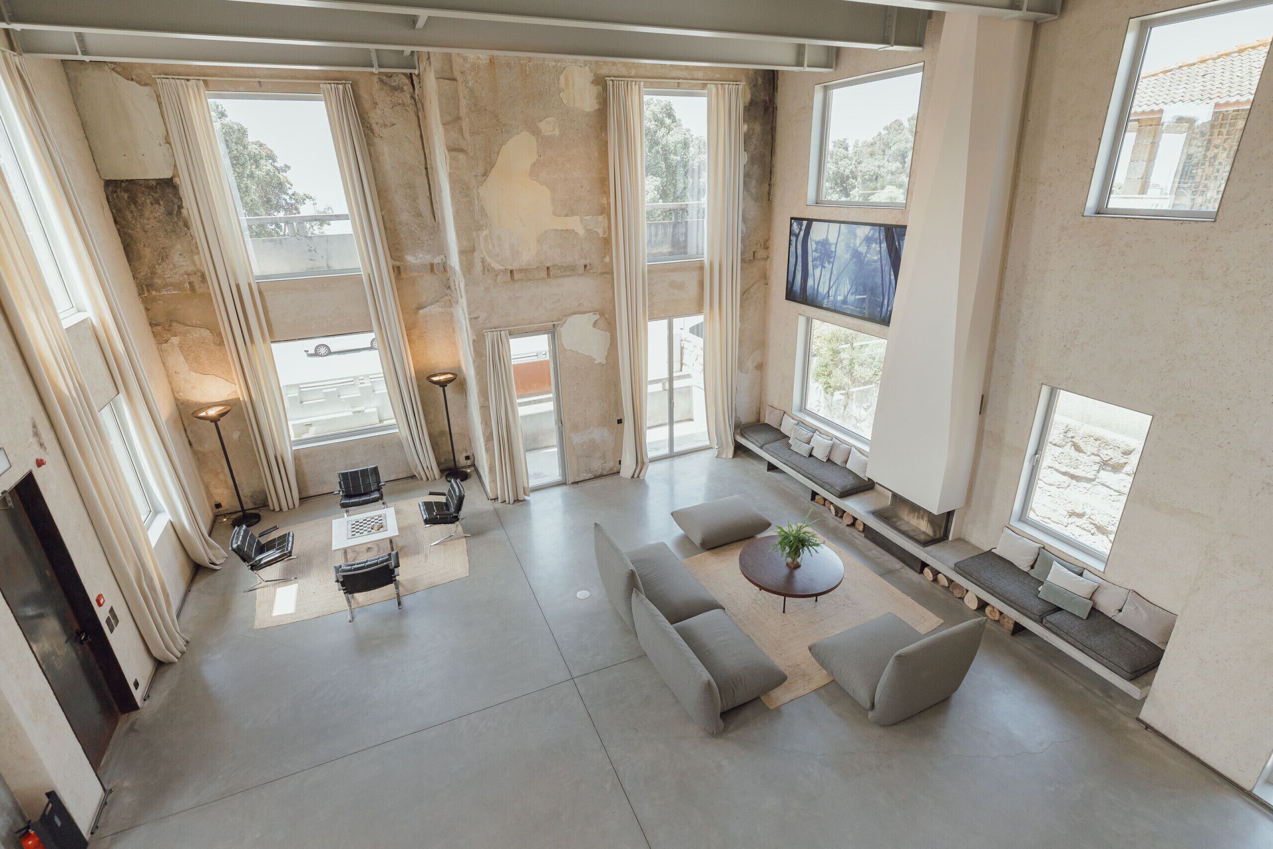 A spacious living room with a large sofa, a coffee table, and a TV mounted on the wall. There are multiple windows providing natural light and views of the outside.