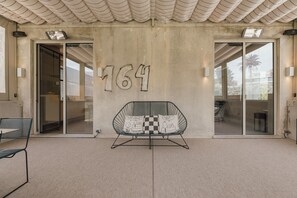 Ein moderner Hauseingang mit der Hausnummer 164 an der Wand, einer Glastür und einem Metallstuhl mit karierten Kissen.