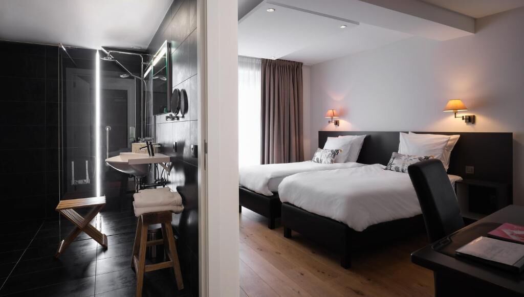 Habitación de hotel moderna con baño privado, cama con dos almohadas, mesita de noche y velador.