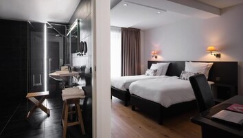 Habitación de hotel moderna con baño privado, cama con dos almohadas, mesita de noche y velador.