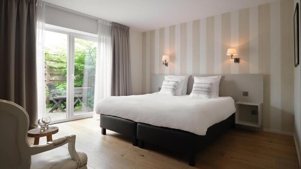 A bedroom with a large bed, two bedside tables, a chair, and a view of the outdoors. - Hôtel Le 24 (Spa)