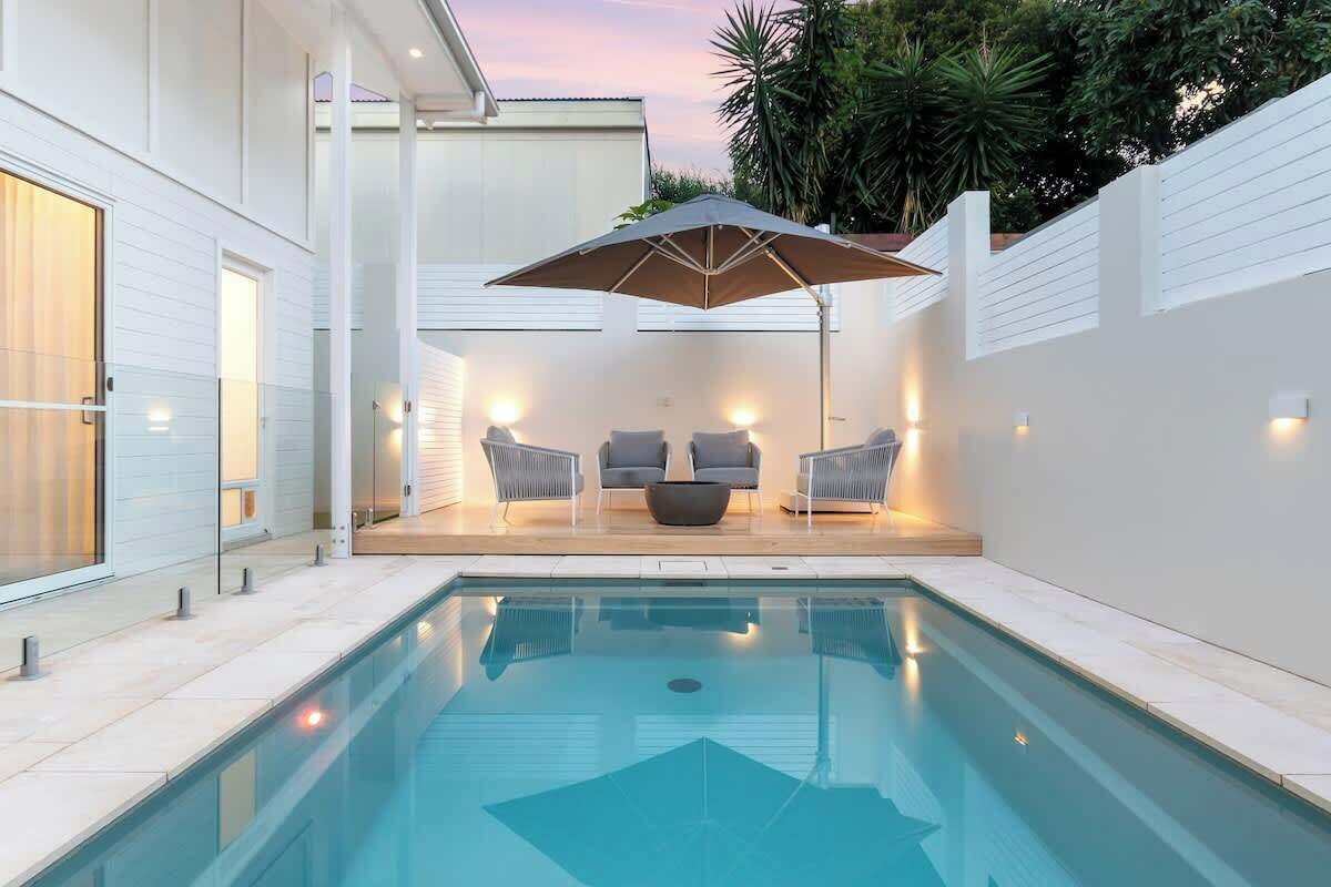 A swimming pool with a sun lounger and two chairs, surrounded by a white wall and a glass door.