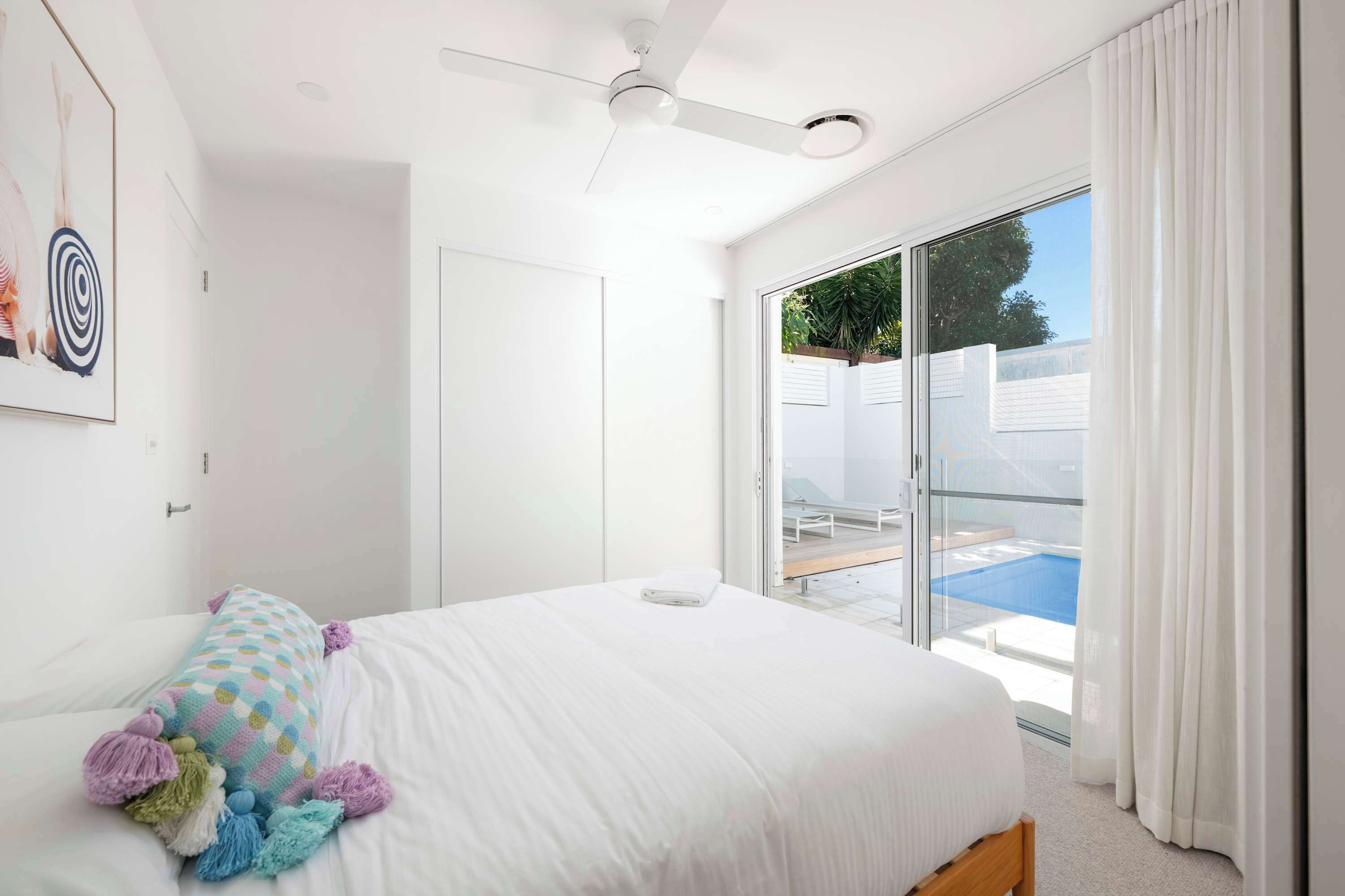 A bedroom with a bed, a ceiling fan, a window with curtains, and a view of a swimming pool.