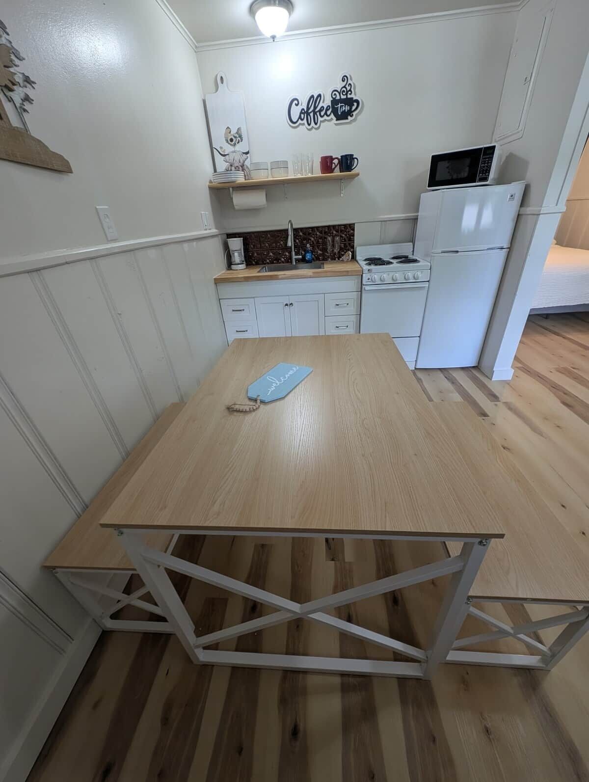 A compact kitchen with a wooden table, white cabinets, a microwave, and a coffee-themed wall decoration.