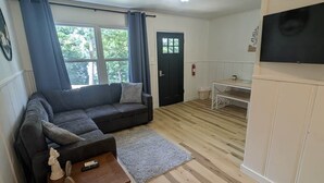 Uma sala de estar com um sofá escuro, uma mesa de madeira pequena e uma TV fixada na parede.