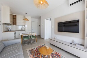Un salon moderne comprenant un canapé, une table basse, un téléviseur fixé au mur et un coin cuisine avec des placards et une table à manger.