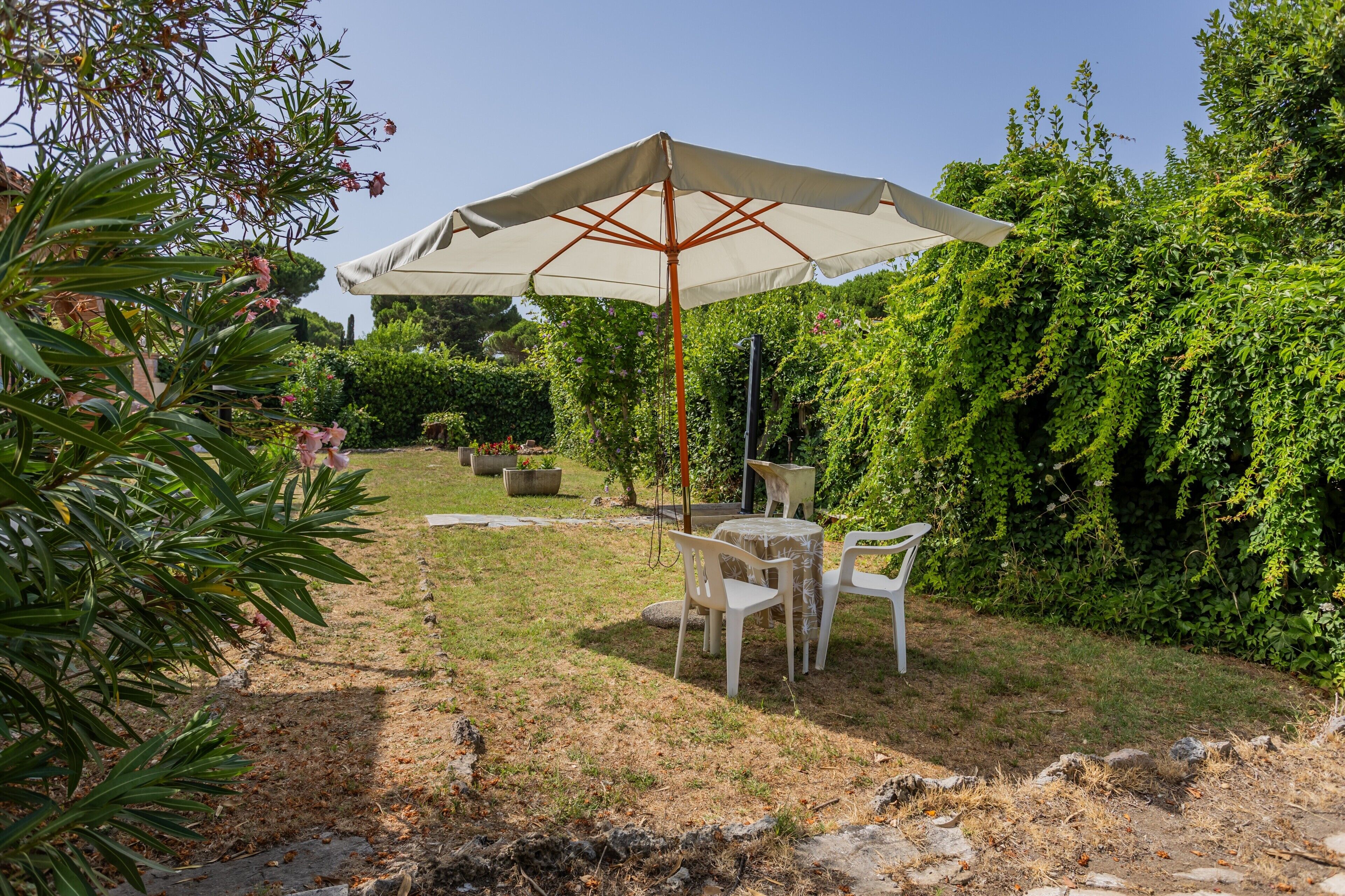Un espace extérieur aménagé avec une table et des chaises sous un grand parasol, entouré d’une végétation luxuriante et d’un jardin bien entretenu.