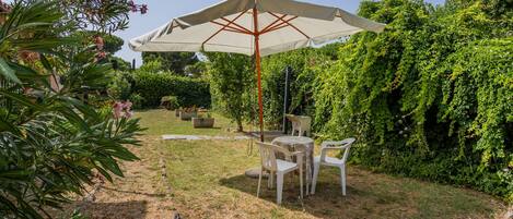 Un espace extérieur aménagé avec une table et des chaises sous un grand parasol, entouré d’une végétation luxuriante et d’un jardin bien entretenu.