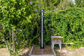 Un jardín con una estructura metálica alta de color negro, un banco de piedra y abundante follaje verde.