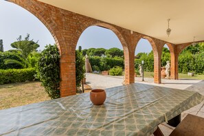Un espacio al aire libre techado, con una mesa y sillas, rodeado de arcos de ladrillo y vegetación.