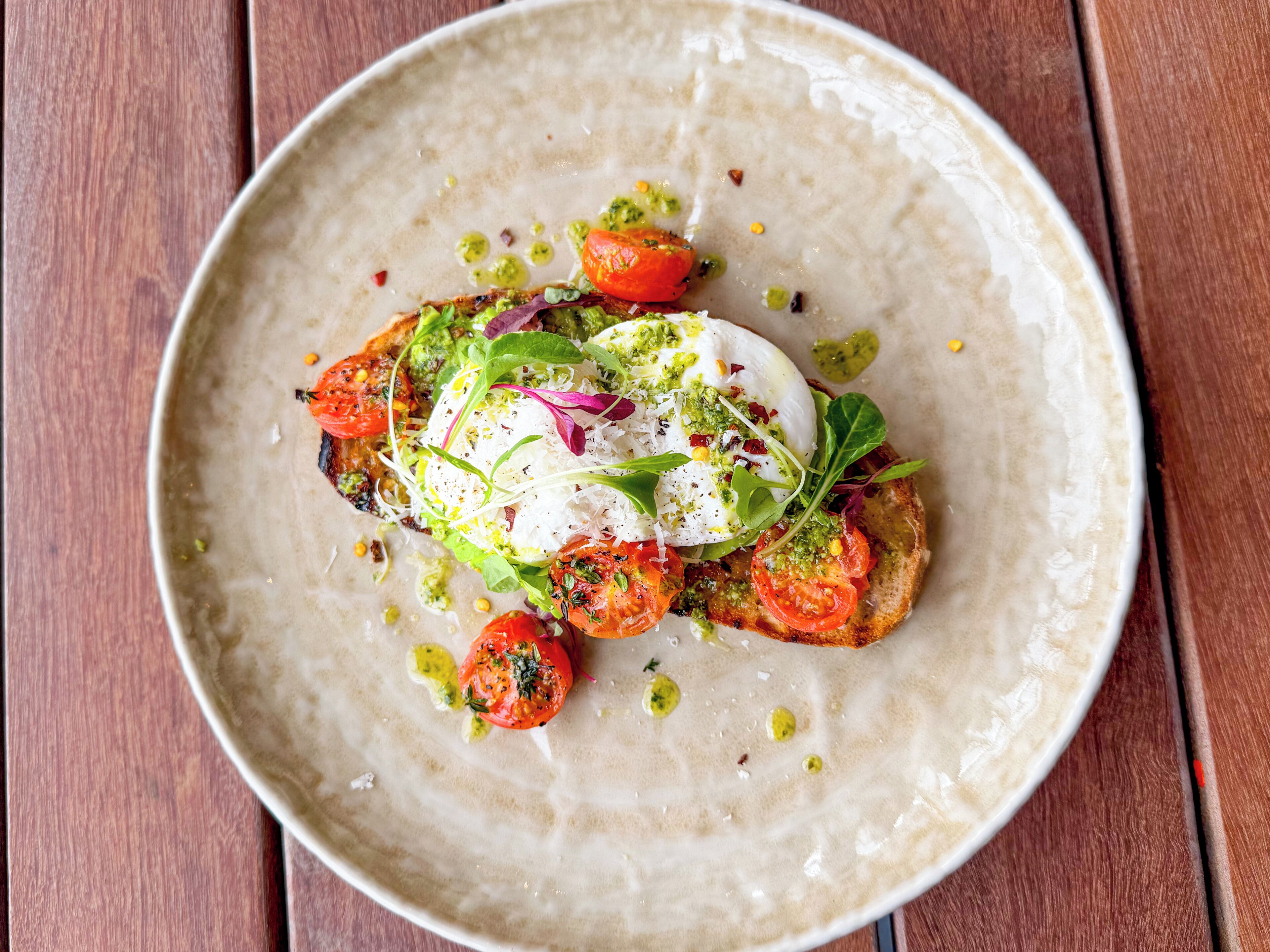 Ein Teller Essen mit pochiertem Ei, Tomaten, Grünzeug und Pesto auf einer Keramikplatte.