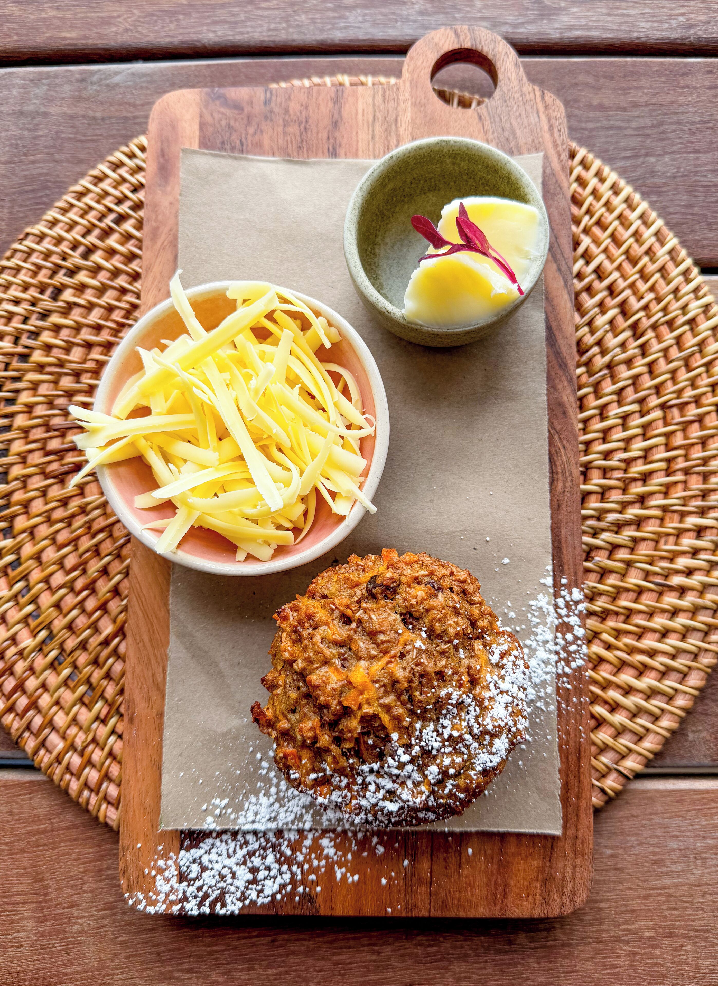 Una tabla de madera con un producto horneado, queso rallado y un tazón pequeño de mantequilla sobre un tapete tejido.