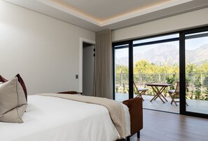 Un dormitorio con una cama grande, vistas a un balcón con mobiliario exterior y montañas al fondo.