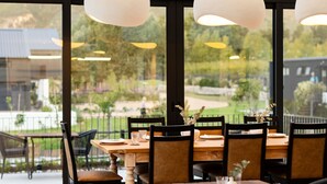 Een restaurant met een groot raam en uitzicht op bergen, houten tafels en zwarte stoelen.