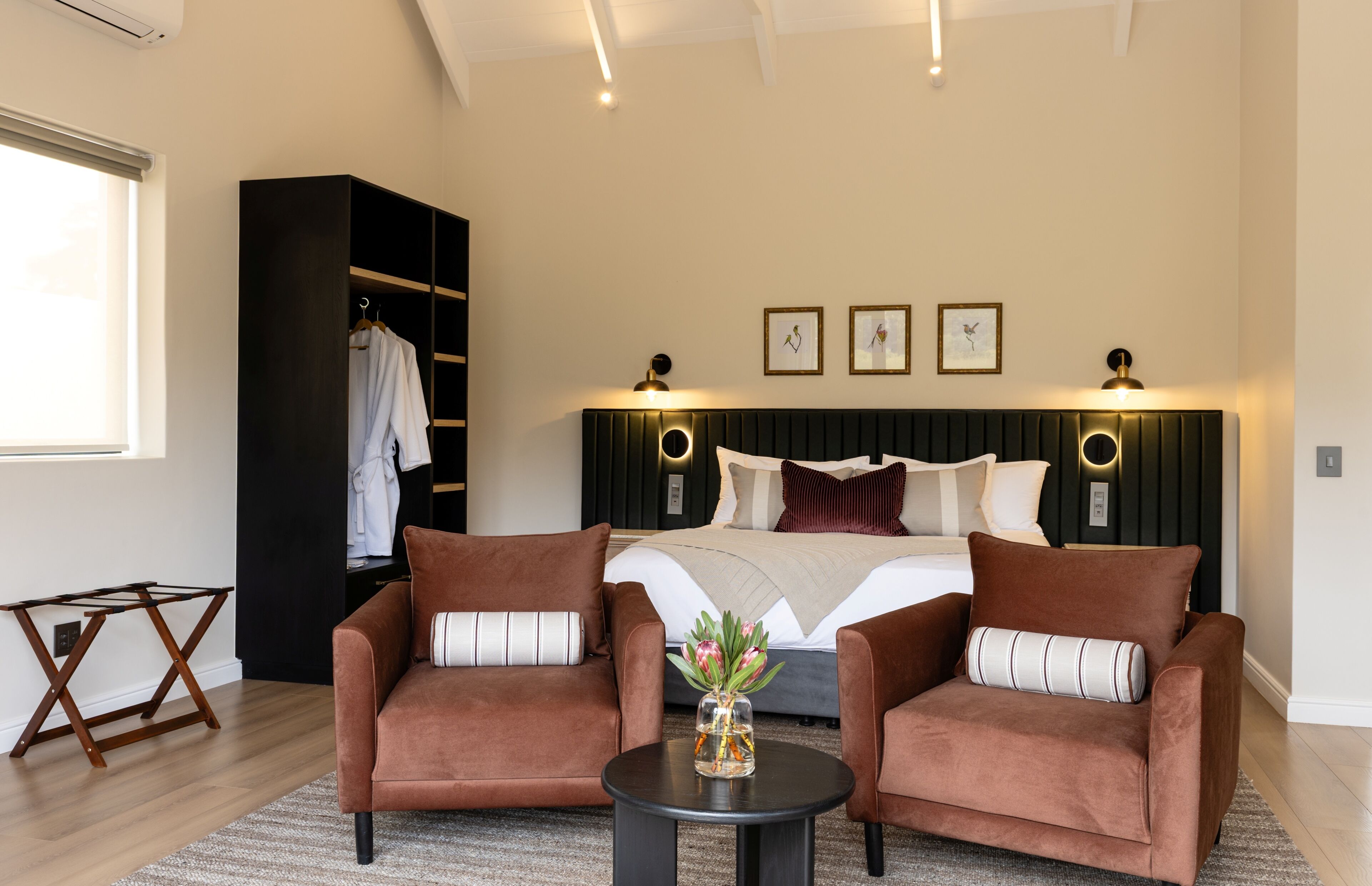 A bedroom with a bed, two armchairs, a small table, a wardrobe, and wall-mounted lights.