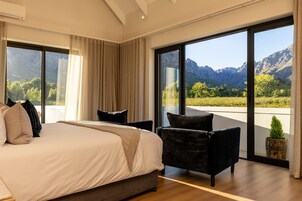 Un dormitorio con una cama grande, dos sillas, vistas a montañas a través de ventanales del piso al techo, y un balcón con planta en maceta.