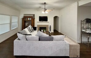 A living room with a grey sofa, a fireplace, a television, and a ceiling fan. - This is a spacious house with 5 bedrooms and 2.5 bathrooms. many amenities (Houston)