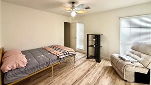 Ein Schlafzimmer mit Bett, Sessel, Deckenventilator, Fenster mit Jalousien und einer Tür.