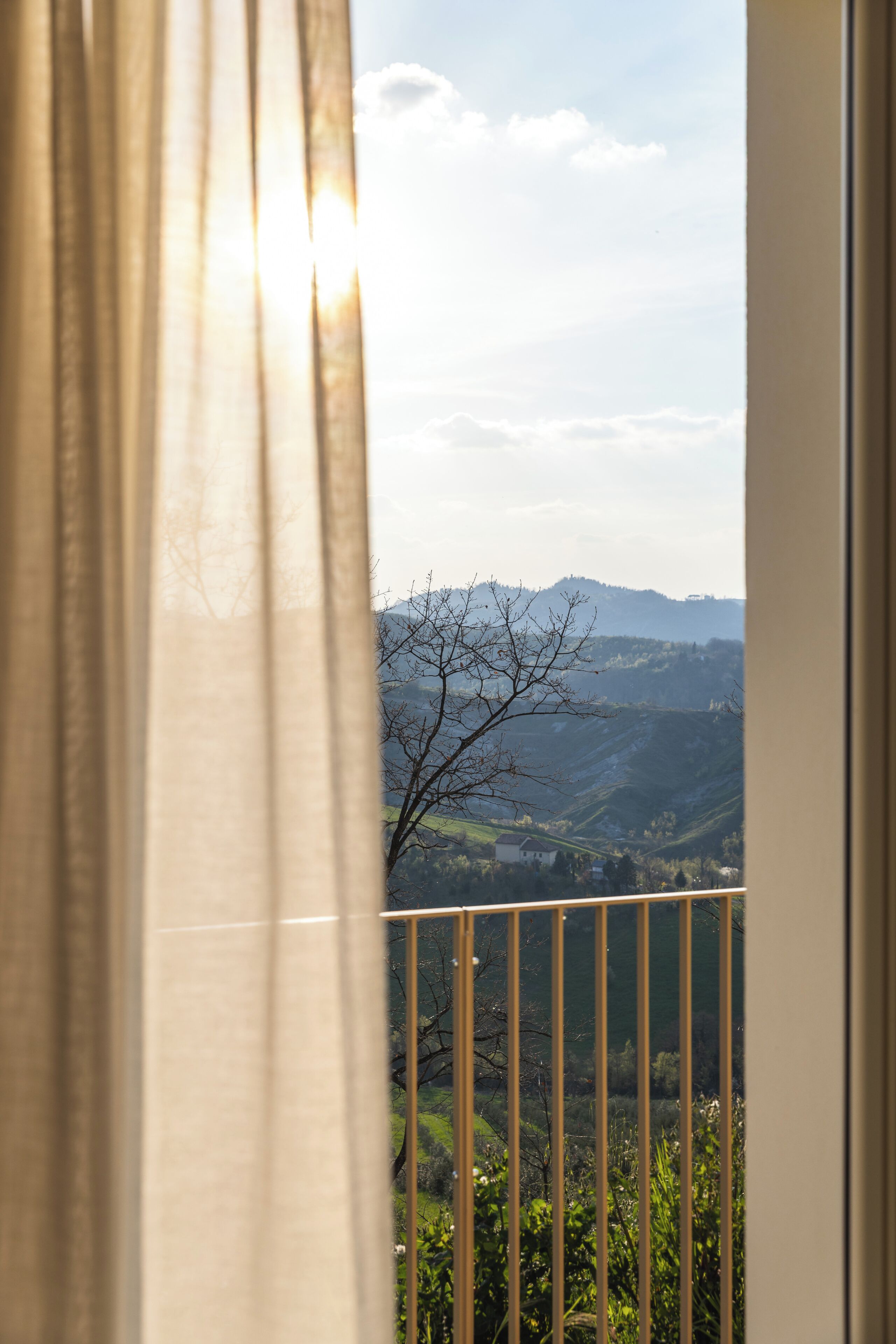 A view through a window with a curtain, overlooking a landscape with hills and a tree.