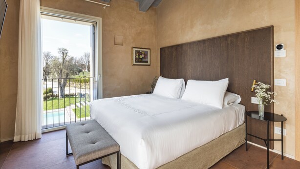 A bedroom with a large bed, a bench, a nightstand with a vase, and a view of a garden through a sliding door.